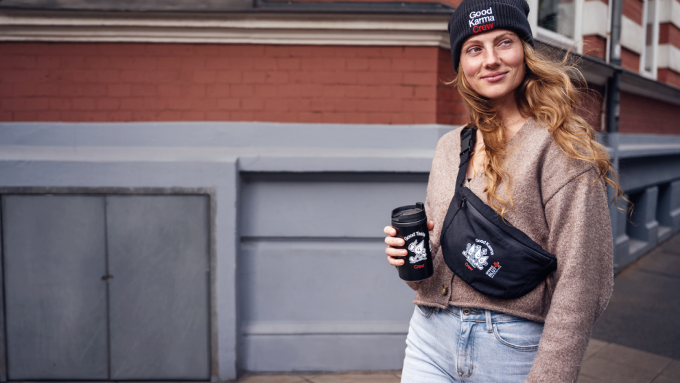 Eine Frau hält einen Kaffeebecher und trägt dazu eine Beanie und eine Bauchtasche.