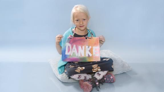 Eva sitzt im Schneidersitz auf dem Boden und hält ein "Danke"-Schild ins Bild