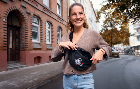 Eine freundlich lächelnde Frau steht mit der Cosy Karma-Bauchtasche in der Stadt