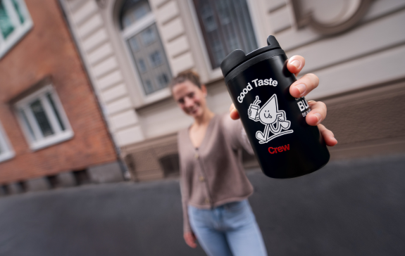Eine freundlich lächelnde Frau hält den Good Taste Crew-Becher in die Kamera