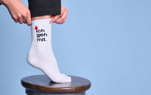 Ein Fuß, der auf einem Hocker steht. Die Person trägt eine weiße Tennissocke mit einem "ich geh mit"-Schriftzug.